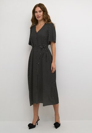 Schwarzes Midi-Kleid mit Pünktchen, V-Ausschnitt, kurzen Ärmeln und einem taillierten Bund. Kombiniert mit schwarzen High Heels mit floralen Akzenten.