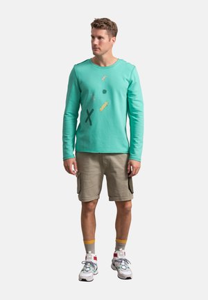 Maglietta turchese a maniche lunghe con vivaci grafiche multicolori, abbinata a pantaloni cargo beige e sneakers bianche con dettagli colorati.