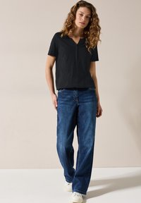 Sort kortærmet top med V-hals og diskrete knapper, parret med løstsiddende blå denimjeans og hvide sneakers. Lys baggrund.