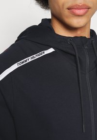 Schwarze Zip-Hoodie mit Kordelzug und einem weißen Akzent. Verfügt über das "TOMMY HILFIGER"-Logo auf der Schulter. Weicher, strukturierter Stoff.