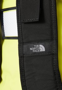 Black fabric strap with a white logo, attached to a bright yellow material with a textured surface and a mesh-like panel.