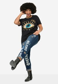 T-shirt nera con una grande grafica di un occhio con dettagli dorati e blu. Jeans blu con scritte bianche e stelle. Stivaletti neri alla caviglia con borchie.