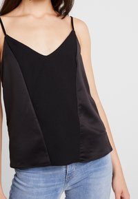 Top noir sans manches avec un décolleté en V, featuring des panneaux mats et satinés, et une texture lisse. Associé à un denim bleu clair.