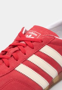 Röd mocka sportskor med vita diagonala ränder och snörning. Har texturerad sömnad och en vadderad krage. Logotyp synlig på tungan.