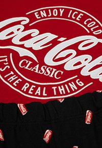 Camiseta roja con el logotipo y eslogan de Coca-Cola en blanco, combinada con pantalones cortos negros con un pequeño patrón de latas de Coca-Cola rojas en la cintura.