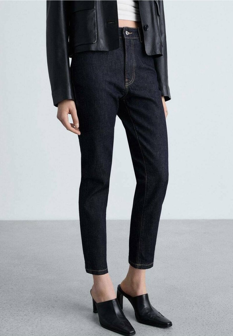 Personne portant un jean bleu foncé, une veste en cuir noire, des mules noires à talons et un haut blanc, debout sur un sol gris uni.