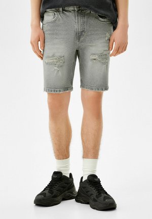 Zblakłe szare szorty dżinsowe z przetartymi elementami, postrzępionymi nogawkami, pięcioma kieszeniami, noszonym wyglądem, w połączeniu z czarnymi sneakersami i białymi skarpetkami.