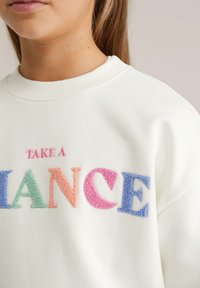 Jauns cilvēks valkā baltu sporta džemperi ar krāsainu teksturētu izšuvumu, uz kura priekšpuses rakstīts "UZŅEMIES RISKU".