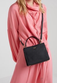 Borsa nera dalla texture strutturata, con una forma definita, dettagli in oro e un manico corto, indossata con una blusa rosa fluente.