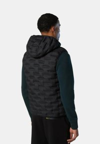 Chaleco negro acolchado con capucha, con textura de acolchado y lados lisos, llevado sobre una camiseta de manga larga oscura.