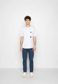 T-shirt bianca a maniche corte con tasca e dettaglio in tessuto nero, abbinata a jeans blu e sneakers bianche, in piedi su uno sfondo neutro.