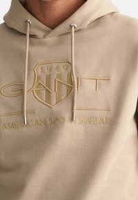 Sudadera con capucha beige con tela texturizada, cordones ajustables y logotipo de la marca en relieve con un diseño de escudo y texto "AMERICAN SPORTSWEAR".