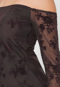 Off-the-shoulder kjole med gjennomsiktige lange ermer, med en brodert blomstermønster i mørk burgunder på en solid mørk bakgrunn.