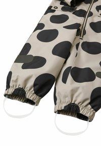 Beige vattentät overall med stora svarta prickar. Har elastiska manschetter och remmar för en säker passform. Slät textur, hållbart material.