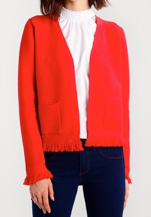 Vrouw draagt een felrode, gestructureerde cardigan met franjes aan de randen, over een witte blouse met hoge hals en donkerblauwe spijkerbroek.