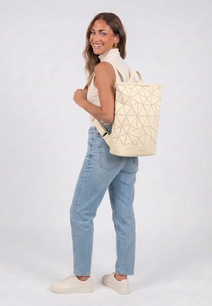 Donna sorridente, indossa jeans azzurri chiari, dolcevita senza maniche color crema, sneakers bianche, porta uno zaino beige con motivo geometrico su una spalla.