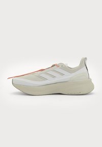 Vaalean beige Adidasin lenkkarit, joissa paksu pohja, valkoiset kolme raitaa sivulla ja oranssit ja valkoiset nauhat, näytetty ulkopuolelta.
