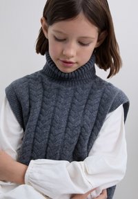 Pige med brunt hår iført en grå kabelstrikket ærmeløs turtleneck over en hvid langærmet bluse, kigger ned med armene korslagte.