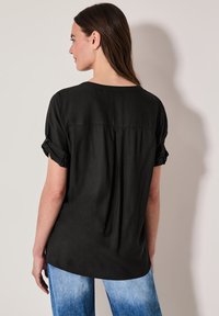 Sort kortærmet top med rullede ærmer, afslappet pasform og en subtil rygsplit, parret med lyseblå jeans. Glat tekstur.