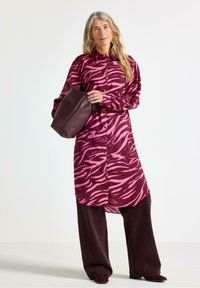 Langærmet skjortekjole med et rosa og bordeaux zebra mønster, knap-front design, og parret med mørk bordeaux brede bukser.