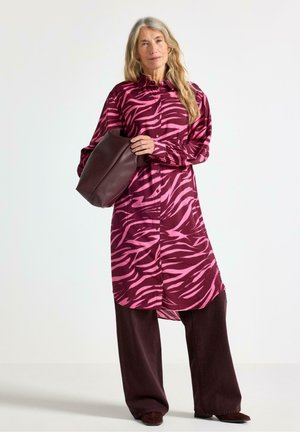 Pitkähihainen mekko, jossa on vaaleanpunainen ja burgundinvärinen seepra-kuvio, napitettava etuosa ja yhdistetty tumman burgundinvärisiin leveylahkeisiin housuihin.