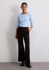 Gestreepte blauw-witte blouse met lange mouwen en een bootnecklijn, gecombineerd met donkere bruine corduroy flared broeken. Model staat tegen een effen achtergrond.