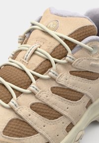 Beige og brune sneakers har en mesh-overdel, rund tå og snøring. Detaljene inkluderer en lys grå krave og teksturerte paneler.