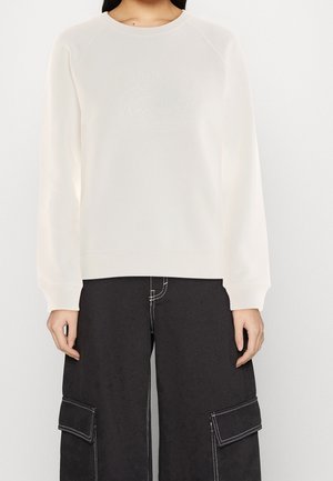 Person trägt einen cremeweißen Langarm-Pullover und schwarze, weit geschnittene Cargo-Hosen mit weißen Nähten und großen Seitentaschen.