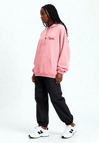 Felpa con cappuccio rosa con la scritta "SWEET heart", grande tasca frontale, abbinata a pantaloni cargo neri e scarpe da ginnastica bianche.