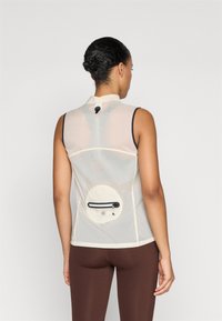 Haut de sport sans manches, de couleur beige clair avec une surface texturée, poche arrière zippée et col haut. Souligné par des bordures sombres.