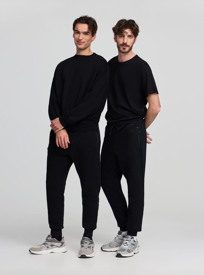 Felpa nera e t-shirt abbinate a pantaloni jogger neri. Tessuto morbido, vestibilità rilassata, tasche con cerniera e stile con sneaker grigie.