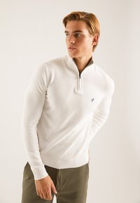 Maglione bianco con zip realizzato in tessuto morbido, con polsini e orlo a coste. Presenta un piccolo logo blu sul petto.