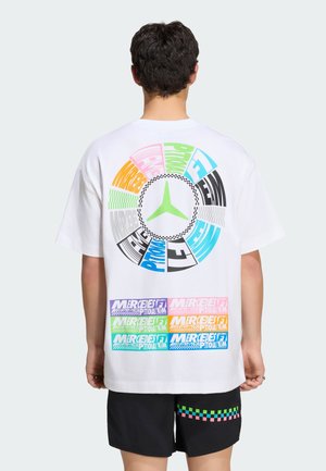 Mężczyzna noszący białą, oversize koszulkę z kolorowym okrągłym emblematem Mercedesa i wielobarwnymi blokami tekstowymi Mercedes F1 na plecach, w połączeniu z czarnymi szortami.