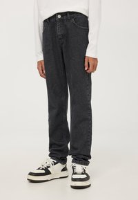 Persona che indossa jeans neri a gamba dritta e sneakers in bianco e nero, in piedi contro uno sfondo chiaro e uniforme.