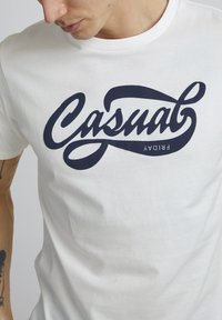 Balta medvilninė marškinė su dideliu marine mėlynu užrašu "Casual" kalligrafiniu šriftu per krūtinę, o žodis "Friday" po juo.