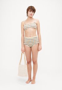 Zweiteiliges Bikini mit kariertem Muster in Pastellfarben, mit einem Bandeau-Oberteil und hochgeschnittenem Unterteil. Model hält eine geflochtene Tragetasche.