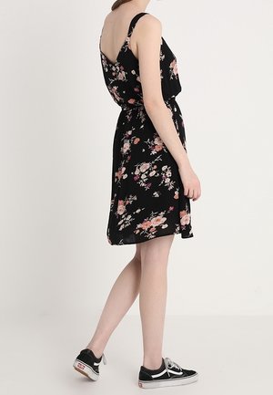 Femme en robe noire fleurie sans manches et baskets noires, debout de profil devant un fond clair uni.