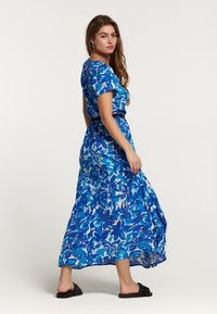 Maksij dress v modri barvi s cvetličnim vzorcem, kratkimi rokavi, prilegajočim pasom in puhastim krilom. Nosi se z črnimi sandali, kar nudi lahek material.