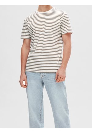 Mand iført en beige og hvid stribet kortærmet t-shirt og lyseblå denim jeans, der står foran en ensfarvet baggrund.