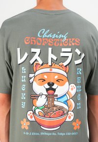 Olijfgroene t-shirt met een cartoon hond die eetstokjes boven een ramenkom houdt, met vetgedrukte meerkleurige tekst en bloemenaccenten.