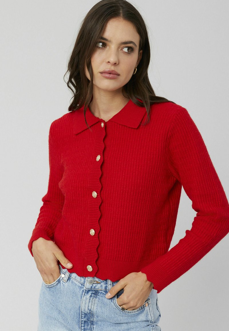 Cardigan rouge côtelé avec un col festonné, des boutons dorés et des manches longues, associé à un denim bleu clair.