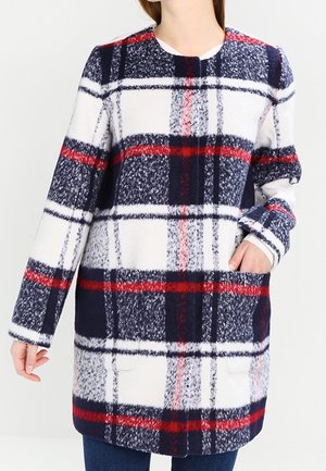 Woman wearing a white, navy, and red plaid long-sleeve coat with front pockets, paired with blue jeans.