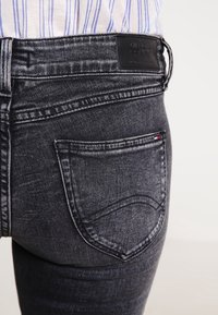 Džínové kalhoty tmavého praní mají mírně poškozenou strukturu, malou zadní kapsu se šitými detaily a pásek s logem.