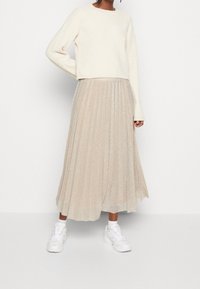 Person in einem cremefarbenen Langarmpullover, beiger plissierter Midirock, weißen Socken und weißen Sneakers, stehend vor einem schlichten weißen Hintergrund.