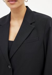 Črn blazer z strukturnim ovratnikom, zapiranjem na en gumb in žepom na prsih. Gladka tkanina z izbranim krojem, primerna za formalno obleko.
