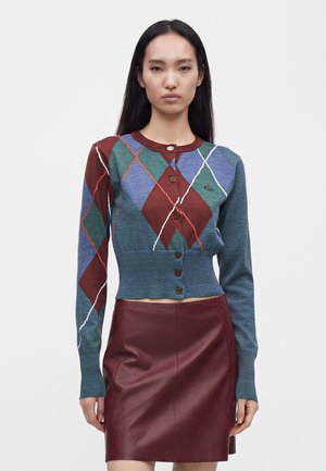 Kvinde iført en tætsiddende argyle mønstret knappet cardigan i blå, grøn og burgunder med en burgunderrød lædernederdel mod en ensfarvet baggrund.