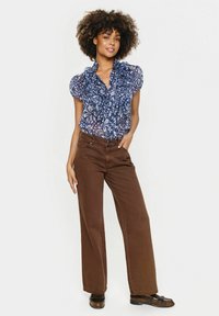 Modra bluzica s cvetnim vzorcem in volanastim ovratnikom, v kombinaciji s širokimi rjavimi hlačami. Model stoji na nevtralni podlagi. Vidna je tudi rjava obutev.