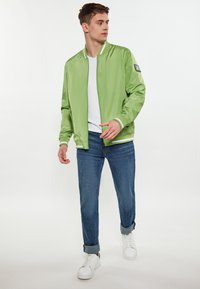 Šviesiai žalia bomber tipo striukė su baltais akcentais, dėvima ant baltos marškinėlių. Derinama su mėlynais džinsais ir baltais sportbačiais. Lygus, blizgus audinys.