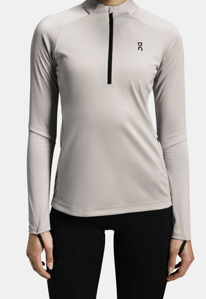 Haut de sport gris clair à manches longues avec fermeture éclair noire jusqu'à la poitrine et petit logo sur la poitrine, porté avec un legging noir.