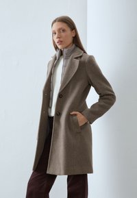 Manteau en laine marron avec un col à revers, doté de deux boutons et de poches latérales. Porté par-dessus une veste matelassée gris clair et un haut blanc.